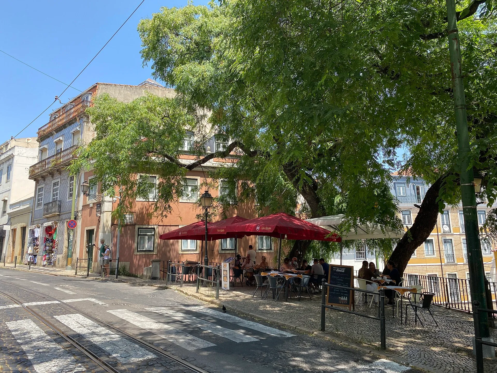 Tables under red parasols in the shade of a tree on a cobbled street in Alfama, tram rails in the foreground and tiled house fronts behind