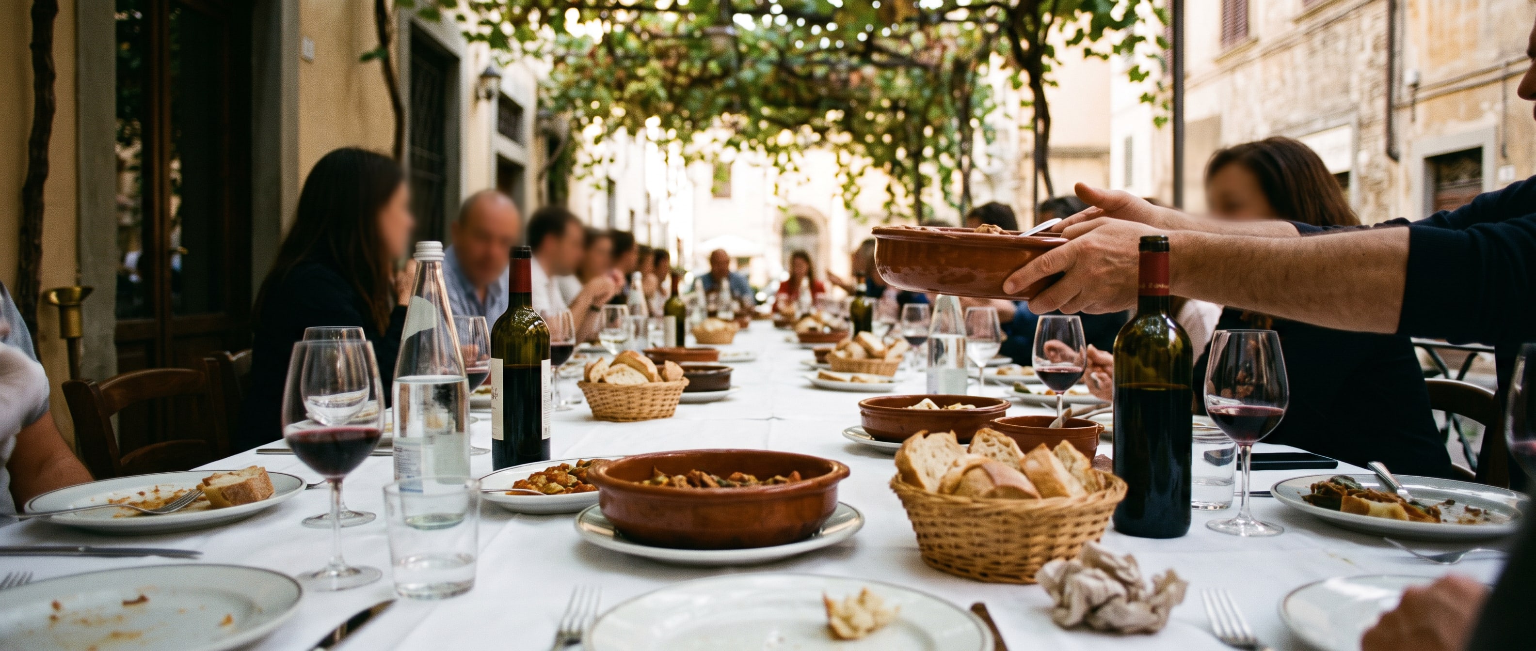 A long shared table at a Sunday lunch seen from one end: plates, bread, bottles and glasses down its length and many hands passing dishes.