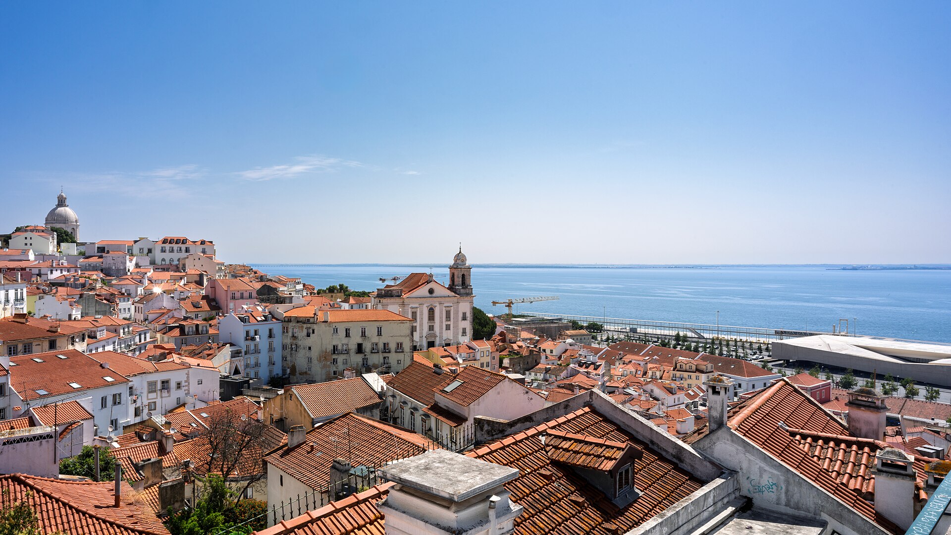 The red-tiled roofs of Alfama stepping down to the wide river Tagus under a clear midday sky, a white church tower and the dome of the National Pantheon above them